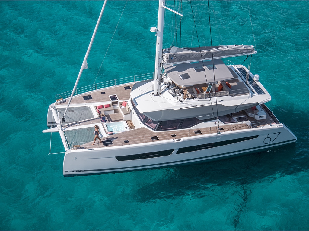 Aerial view of a large, modern catamaran yacht anchored in clear, turquoise waters. The deck offers ample seating and a lounge area, the sails are neatly folded. Visit us at the