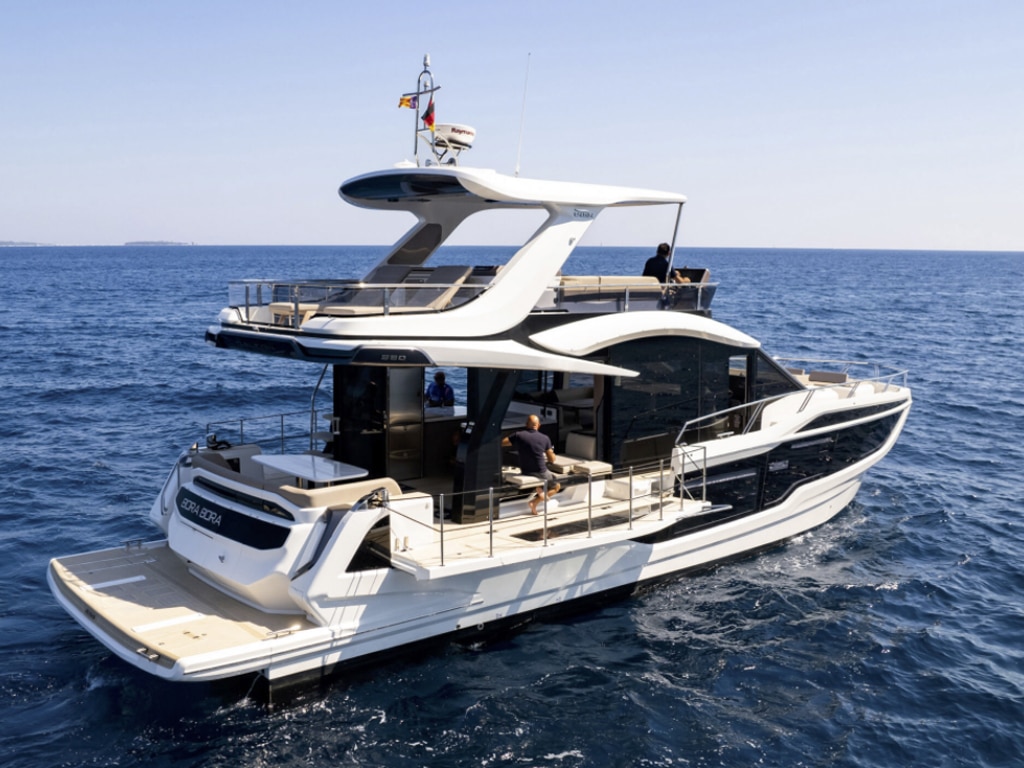 A luxurious white yacht with two decks, cruising on a calm blue sea under clear skies. The yacht has several outdoor seating areas and a rear platform. A Spanish flag flies on the roof and invites you in,