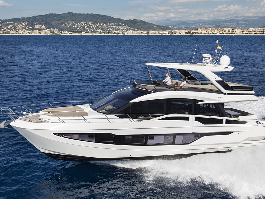 A modern white luxury yacht cruises on the blue sea, with a clear sky and a coastal town in the background. Visit us at the Palma International Boat Show.