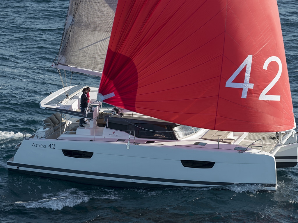 A white sailing yacht with a large red sail bearing the inscription "42" glides through the water, with a man at the helm steering the ship. Visit us at the Palma International Boat