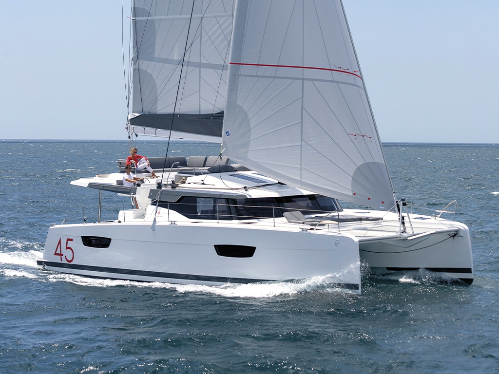 A modern catamaran with white sails, marked with the number 45, sails on a calm blue ocean. Two people can be seen on the deck, one is steering behind. Visit